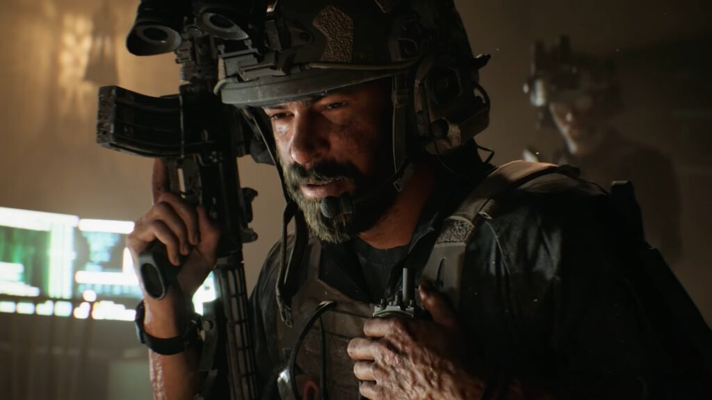A soldier grips an assault rifle. Another can be seen behind him, wearing a helmet and binoculars