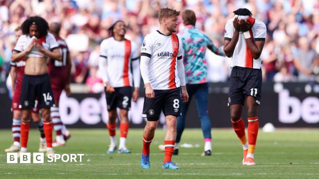 Luton Town players Luke Berry and Elijah Adebayo in white jerseys with orange and black stripes appear dejected on the pitch, with other players and spectators in the background. Adebayo has his shirt pulled up over his face.
