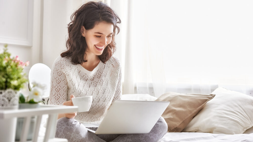 Happy woman sitting on a bed with a coffee and a Windows 10 laptop