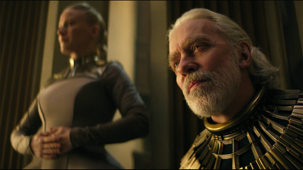 Brother Dusk sitting down with an out-of-focus Demerzel nearby in Foundation season 3 episode 8
