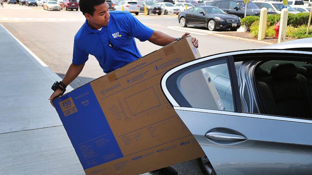 A Best Buy employee loads a boxed television into a silver car on a sunny day.