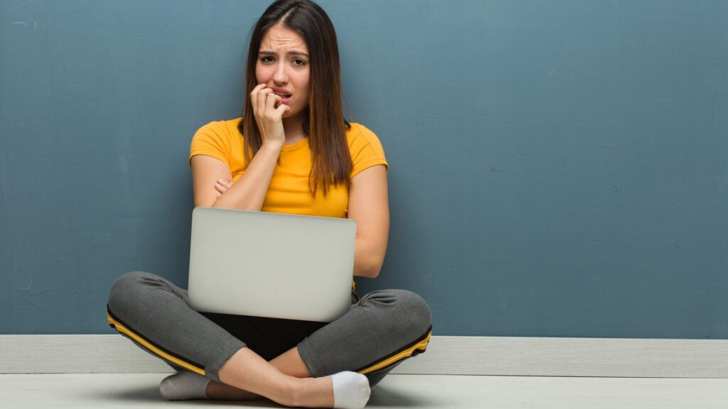 Young woman sitting on the floor with a Windows 10 laptop biting nails, nervous and very anxious